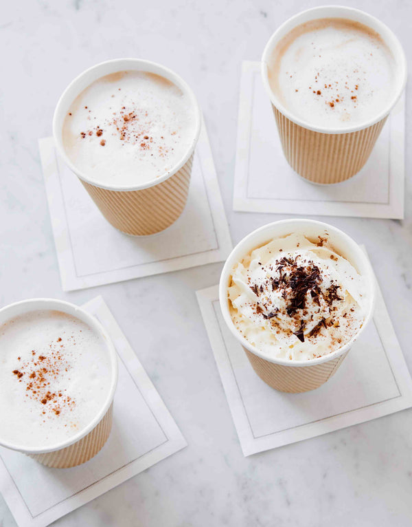 Photo of four coffees, lattes, and cappuccino drinks in kraft cups. Topped with whipped cream, cocoa powder, and cinnamon.