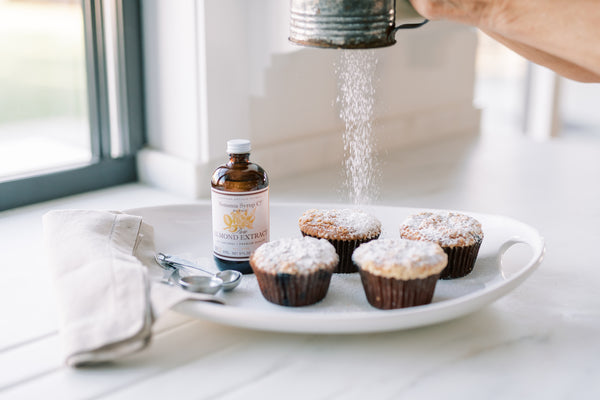 Picture of muffins on plate next to almond extract with powdered sugar falling onto muffins