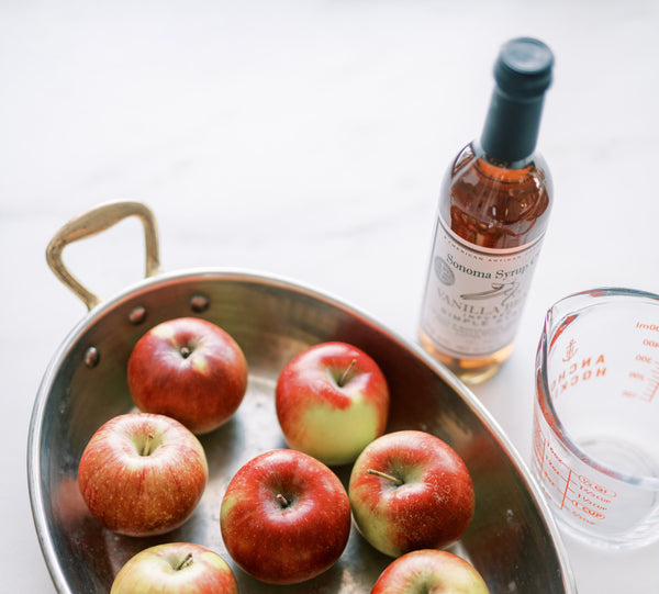 Photo of a large dish full of fresh red apples next to a measuring cup and bottle of Vanilla Bean Simple Syrup.