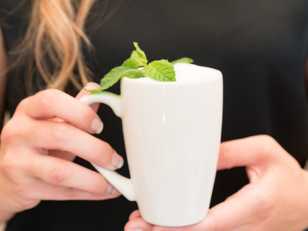 Picture of women holding tall white mug full of mocha topped with whipped cream and sprigs of peppermint leaves