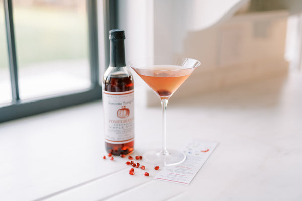 Photo of a Pomegranate Martini which has a slight pink color and cherry at bottom of glass. Bottle of Pomegranate Simple Syrup and fresh pomegranate seeds sits next to the martini glass.