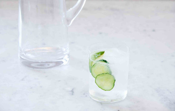 Picture of a clear cocktail with floating cucumber slices next to a large pitcher