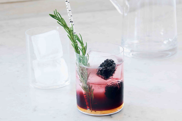 Picture of clear glass filled with dark berry cocktail, fresh ice, black berries, and a sprig of rosemary on a marble table