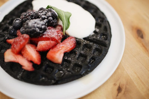 Picture of white plate on table with chocolate brown waffle topped with whipped cream, strawberries, blackberries, blueberries, and mint sprig